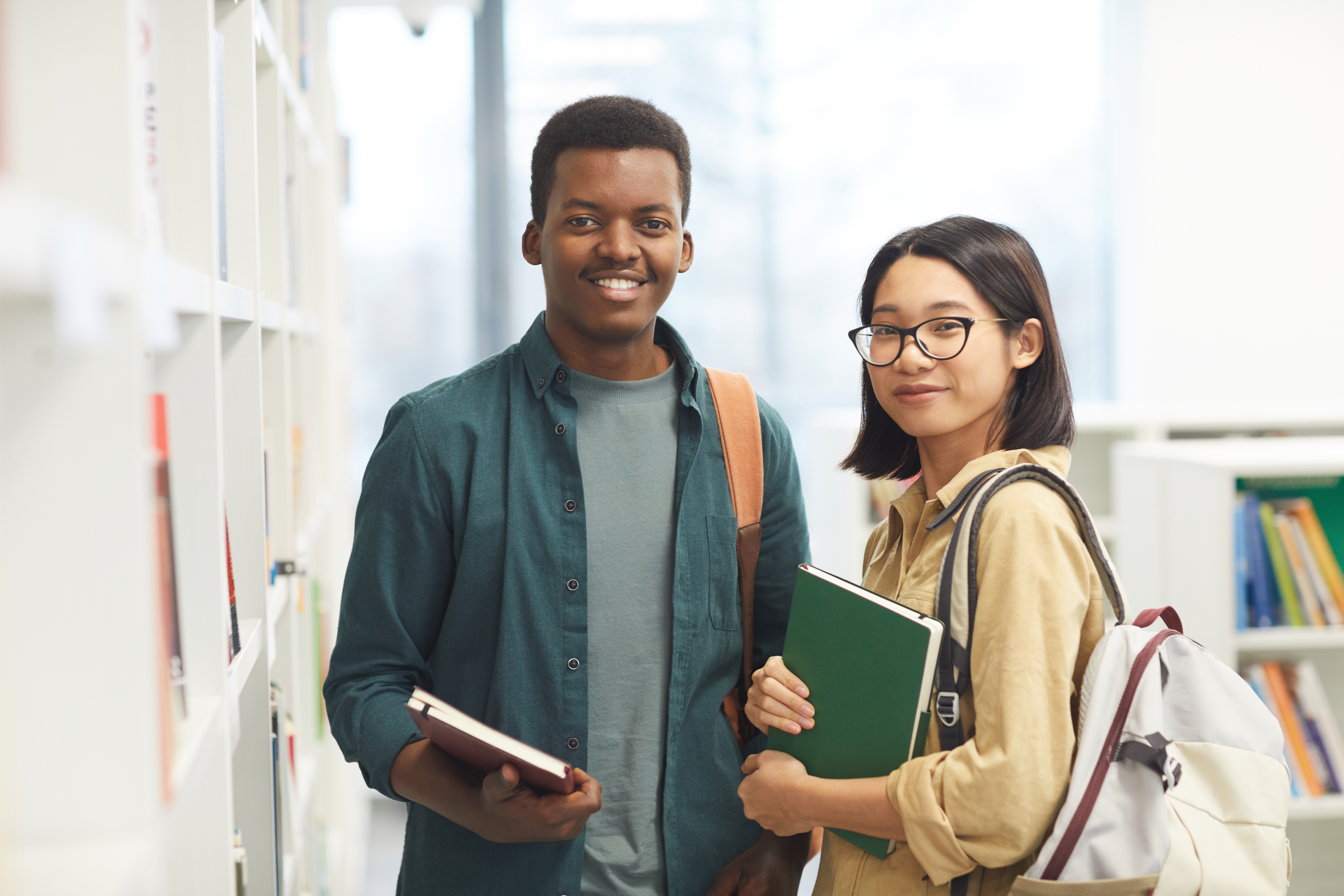 A male student of African descent stands next to a female student of asian descent. Both are holding books with bakcpacks draped over their shoulders.