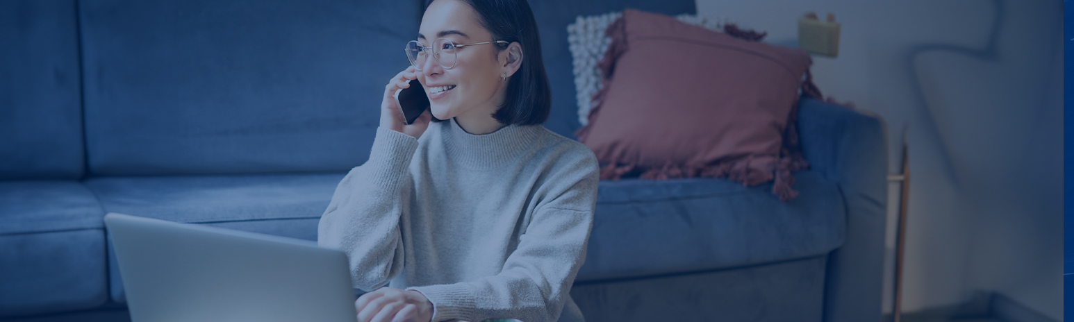 Frau mit schwarzen Haaren und Brille im grau-weißen Pulli hat Handy in rechter Hand und Laptop vor sich stehen. Sie sitzt auf dem Boden vor einer Couch mit Kissen in der rechten Ecke. Das Bild hat einen blauen Filter.