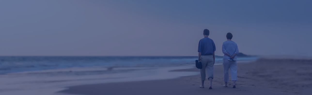 Ein älteres Paar läuft im rechten Bildrand an einem Strand entlang. Beide haben ihre Schuhe ind er Hand und sind nur von hinten zu sehen. Über dem Bild liegt ein blauer Filter.