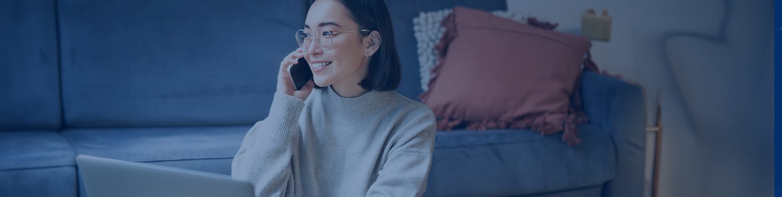 Frau mit schwarzen Haaren, Brille und grau-weißem Pullover sitzt mit Handy in der rechten Hand und Laptop vor sich vor einer Couch. Das Bild hat einen blauen Filter.