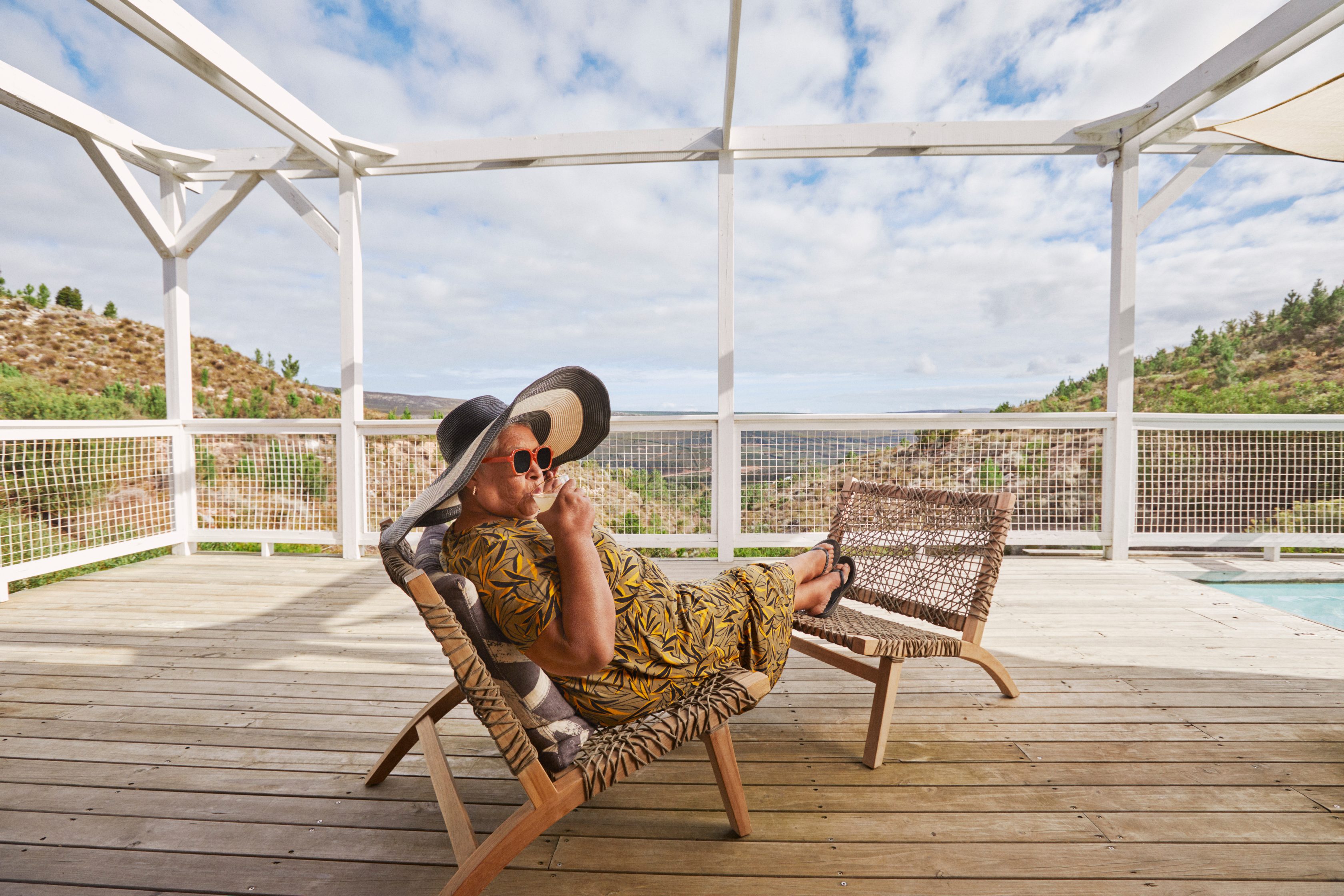 Eine Frau mit Sonnenhut und Sonnenbrille sitzt auf einem Stuhl, legt ihre Füße hoch und trinkt ein kaltes Getränk. Sie sitzt an einem Pool und im Hintergrund ist eine weite Berglandschaft und ein bewölkter Himmel zu sehen.