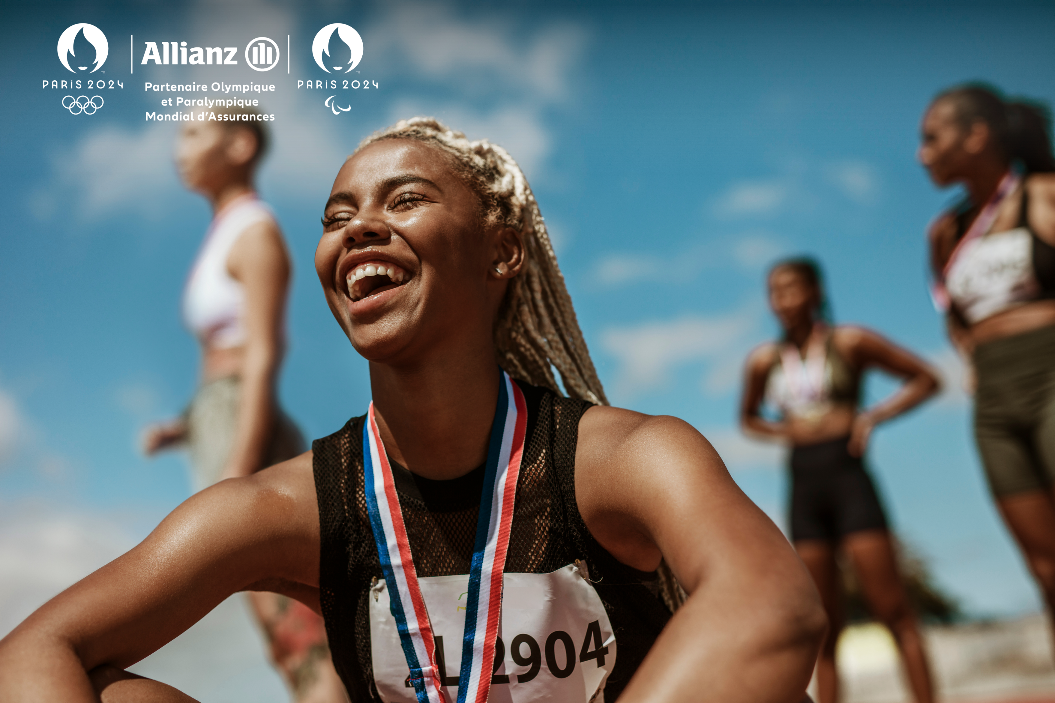 Black female athlete laughing sitting with a ribbon around her neck, surrounded by other athletes blurred in the background. 